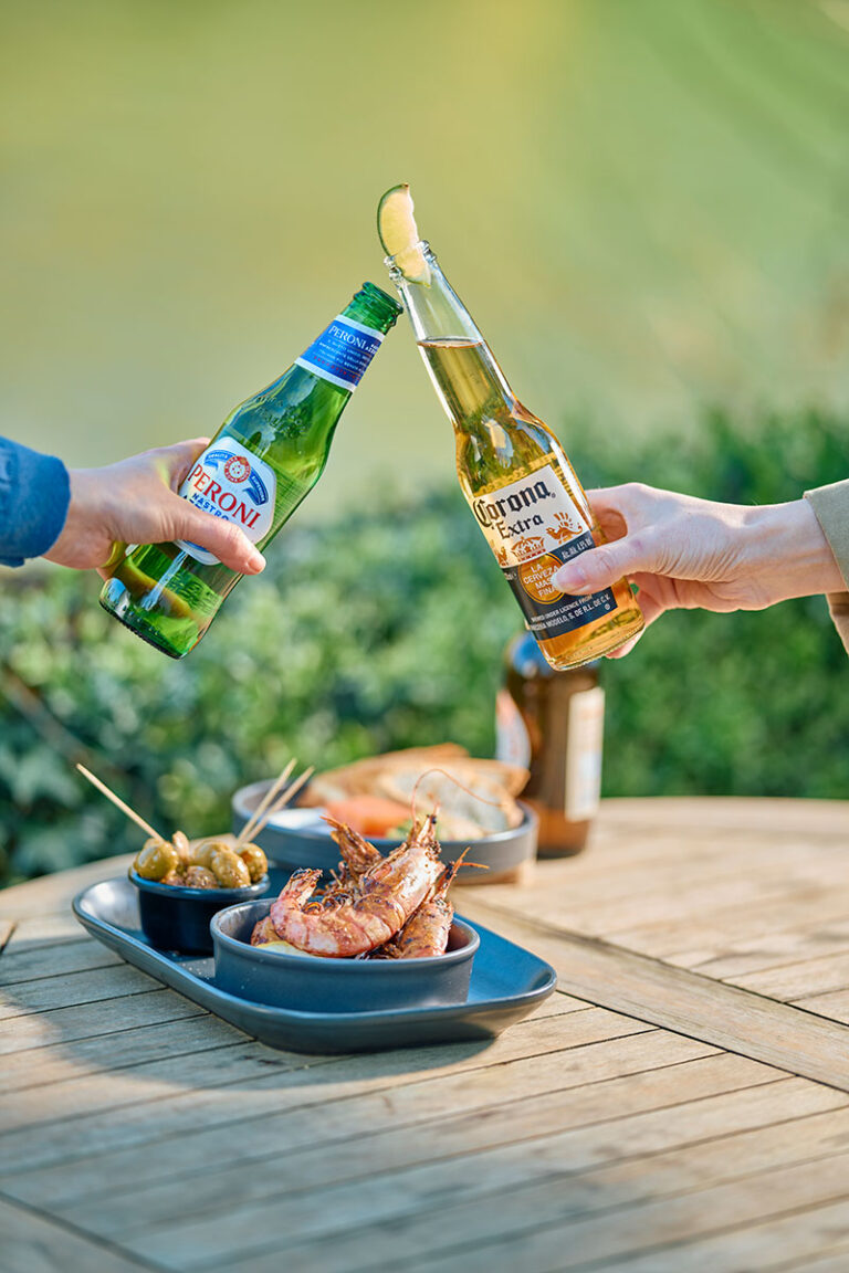 Two bottles of beer held in a cheers toast. Large prawns in a bowl on a wooden table.