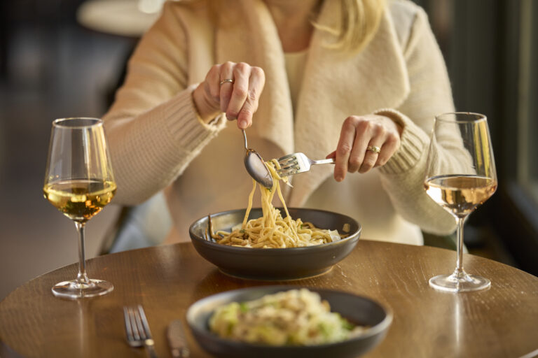 Person sat twirling pasta with a fork and spoon. in the foreground is another plate of food in soft focus.
